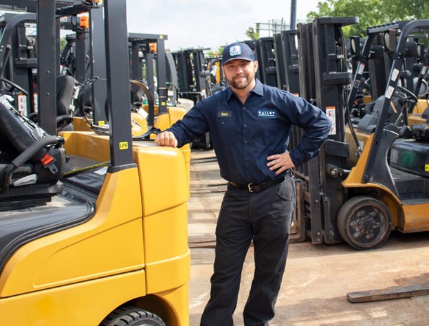 Person wearing a dark Bailey-branded uniform and cap standing outdoors in a lot filled with multiple forklifts. The individual is leaning on a yellow forklift, with several other forklifts lined up in the background.