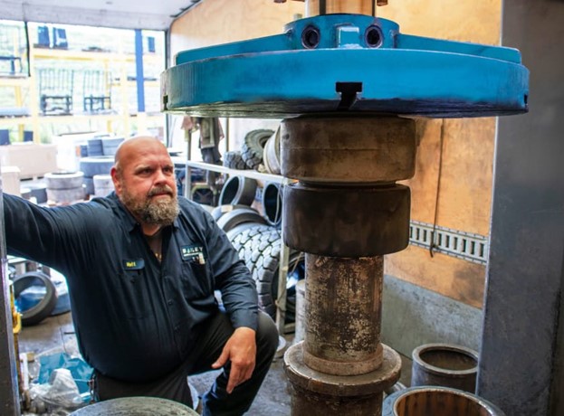 Person wearing a dark work uniform with a Bailey logo kneeling next to a large industrial machine in a workshop. The machine has a blue top component and stacked cylindrical parts, with various tires and metal components visible in the background.