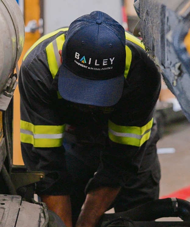 Person wearing a navy Bailey cap and dark work uniform with high-visibility yellow reflective stripes performing maintenance on industrial equipment inside a workshop.