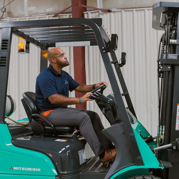 Person operating a green Mitsubishi forklift inside an industrial facility. The forklift has a protective overhead guard and visible hydraulic components, and the background shows a corrugated metal wall and structural beams.