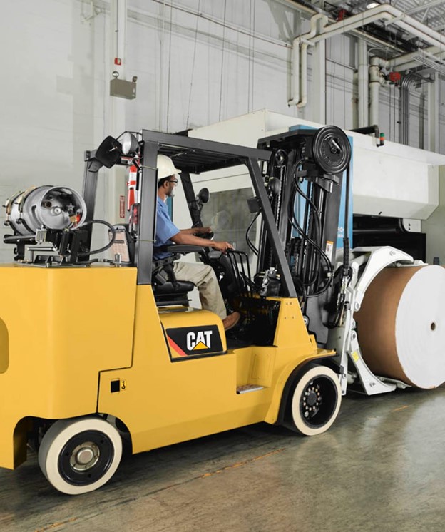 A person operating a yellow CAT forklift inside an industrial facility, lifting a large cylindrical roll of paper using a specialized clamp attachment.