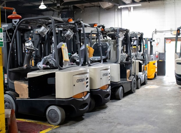 Several Crown brand forklifts lined up inside an industrial warehouse, with a yellow forklift visible in the background. The forklifts appear to be new or recently serviced, and the area has concrete flooring and overhead lighting.